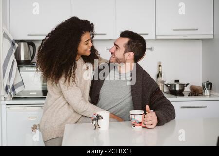 Belle femme afro-américaine souriante et accueillante homme caucasien charmant tout en passant du temps dans une cuisine confortable le matin Banque D'Images