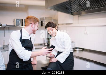 De dessous de la photo de deux gars en uniforme de cuisinier debout sur la cuisine du restaurant et sur les smartphones pendant la pause Banque D'Images