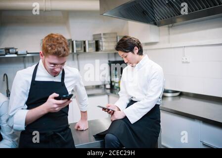 De dessous de la photo de deux gars en uniforme de cuisinier debout sur la cuisine du restaurant et sur les smartphones pendant la pause Banque D'Images