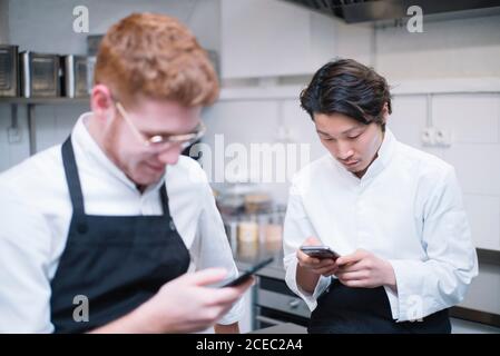 De dessous de la photo de deux gars en uniforme de cuisinier debout sur la cuisine du restaurant et sur les smartphones pendant la pause Banque D'Images