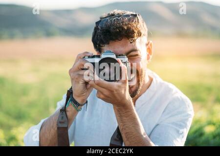 Beau jeune gars en lunettes de soleil souriant gaiement et tenant rétro appareil photo tout en restant debout sur un arrière-plan flou d'une campagne incroyable Banque D'Images