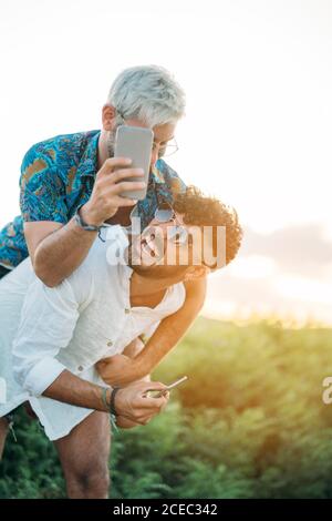 Beau gars barbu donnant le pigeyback à l'ami gai et poser pour le selfie tout en passant du temps dans la nature ensemble Banque D'Images