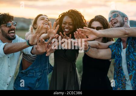 Groupe de jeunes amis divers souriant et se faisant des mains vers l'appareil photo tout en se tenant sur un arrière-plan flou d'une campagne incroyable au coucher du soleil Banque D'Images