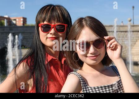 Deux jolies femmes asiatiques dans des lunettes de soleil stylées sourire et regarder à l'appareil photo tout en se tenant près de la fontaine le jour ensoleillé à l'intérieur stationnement Banque D'Images