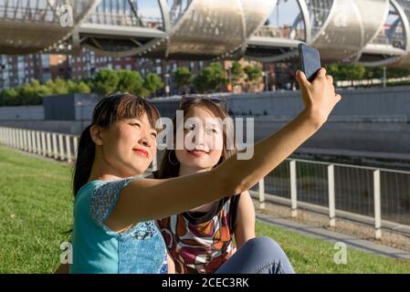 Deux belles femmes asiatiques souriantes et posant pour selfie pendant assis sur le sol par beau temps dans le parc Banque D'Images