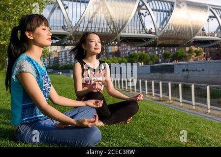 Deux charmantes dames asiatiques garent les yeux fermés et s'asseyant sur le parc herbe dans la pose de lotus Banque D'Images