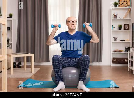 Homme âgé avec vitalité s'exerçant dans la salle de séjour. Personne âgée pensionné en ligne entraînement à l'exercice sur Internet à la maison activité sportive avec haltères, bande de résistance, ballon suisse à l'âge de la retraite. Banque D'Images