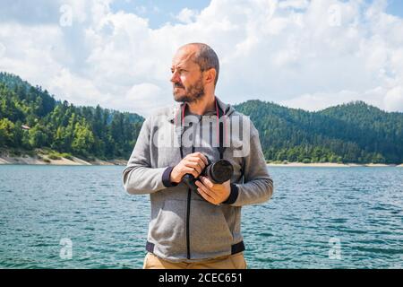 Photographe homme à capuche marchant autour du lac avec un appareil photo reflex numérique et la prise de vue de la nature, portrait à moitié de corps, concept de photographie de paysage Banque D'Images