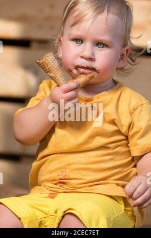 Jeune garçon en vêtements jaunes qui mange de la glace au chocolat avec un cône gaufré. Banque D'Images