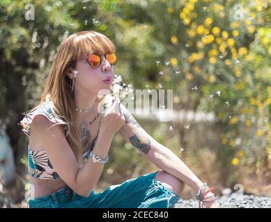 Jeune femme tatouée dans des lunettes de soleil et jeans déchirés assis et soufflant sur un pissenlit blanc moelleux sur un fond de parc flou Banque D'Images