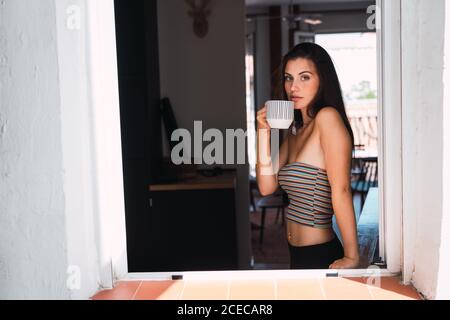 Vue latérale de la jeune belle brune femme en petit top et collants noirs debout près de la fenêtre et boire du café sur le fond de la pièce Banque D'Images