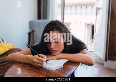 Jeune femme brune en T-shirt noir assise dans la chambre près d'une table en bois et écrivant dans un carnet ouvert Banque D'Images