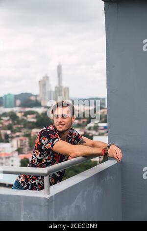 Jeune homme attrayant souriant et penché dans une barre d'acier sur terrasse dans la ville moderne et ciel nuageux Banque D'Images