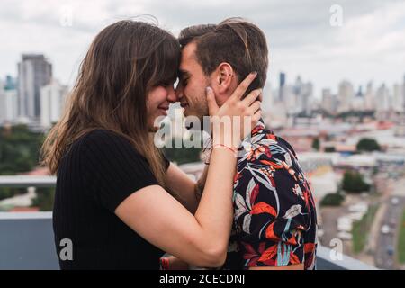 Vue latérale d'un jeune homme et d'une jeune femme souriant et touchant le nez tout en se tenant sur la terrasse sur un fond flou d'une ville moderne merveilleuse Banque D'Images