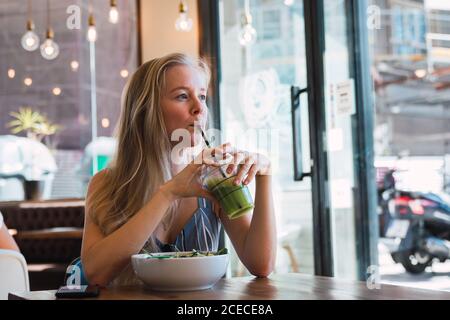 Belle femme blonde qui boit du smoothie vert tout en se refroidissant seul dans un café avec un bol de nourriture à proximité Banque D'Images