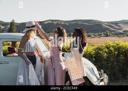 Les femmes multiethniques à la mode se tenant près d'une voiture d'époque et de la carte de lecture lors d'un voyage ensemble en été Banque D'Images