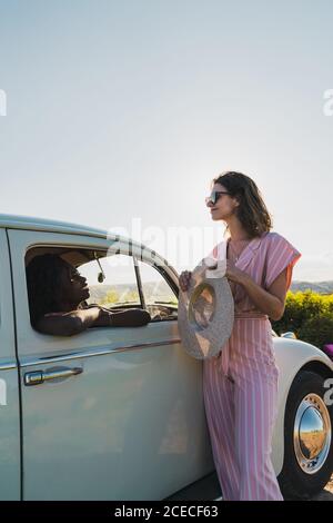 Brunette élégante en lunettes de soleil penchée sur la voiture à l'extérieur et parlant à la jolie femme noire à l'intérieur en plein soleil d'été Banque D'Images