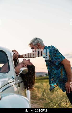 Vue latérale d'une femme assise à l'intérieur d'une voiture penchée tête dehors et regardant un homme debout au-dessus et souriant heureux en été Banque D'Images