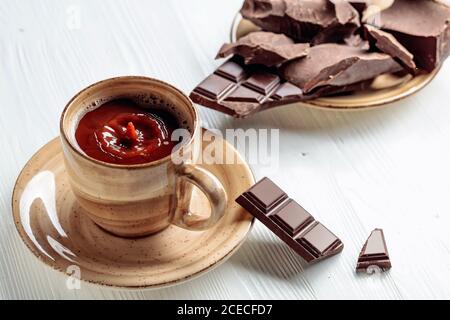 Chocolat chaud et morceaux de chocolat cassés sur une table en bois blanc. Une goutte de boisson tombe dans une tasse. Banque D'Images