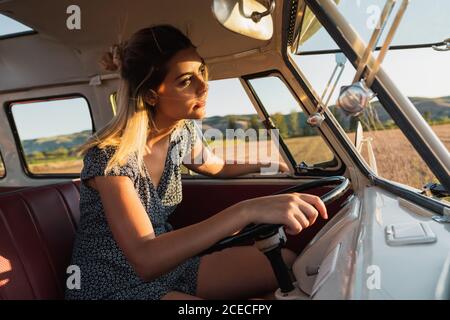 Vue latérale d'une jolie jeune femme assise sur le siège du conducteur et à la conduite d'une fourgonnette d'époque lors d'un voyage dans la campagne sous le soleil jour Banque D'Images