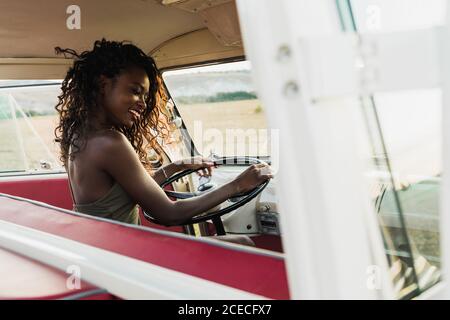 Vue latérale d'une charmante femme afro-américaine souriant et regardant l'appareil photo tout en étant assise sur le siège conducteur d'une voiture rétro et en touchant les cheveux bouclés pendant le voyage en campagne Banque D'Images