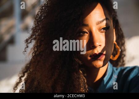 Jeune femme afro-américaine avec des cheveux bouclés et un maquillage éclatant regardant à distance avec l'ombre sur le visage Banque D'Images