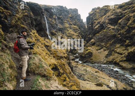 Jeune homme barbu avec sac à dos debout dans les montagnes près de petites cascade et rivière en profitant de la vue et souriant Banque D'Images