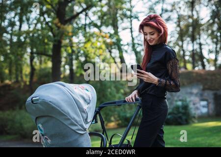 Belle jeune femme avec des cheveux de gingembre souriant et utilisant moderne smartphone tout en marchant avec la poussette de bébé dans le parc Banque D'Images