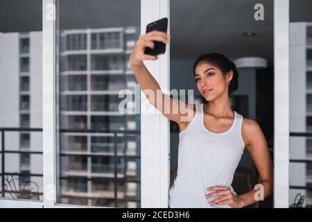 Belle brunette femme hispanique prenant selfie sur téléphone mobile et se tenant entre les fenêtres de balcon Banque D'Images