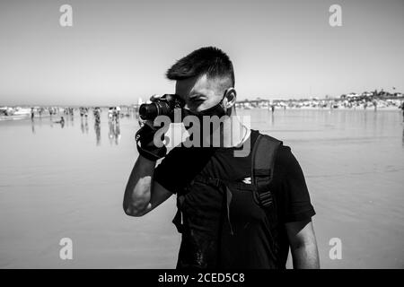 Prise de vue en échelle de gris d'un jeune homme portant un masque médical et prendre une photo à la plage Banque D'Images