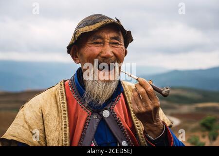 DONGCHUAN RED, YUNNAN, CHINE - 14 JUIN 2018 : homme senior décontracté dans des vêtements traditionnels lumineux assis au sol fumant du tabac en bois et chien pétant assis?à proximité en regardant loin sur un fond flou des terres rouges de Dongchuan dans la province chinoise du Yunnan Banque D'Images