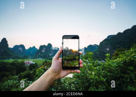 Rognez la main tenant le smartphone en prenant des photos de la belle vallée verte avec des maisons et des arbres entourés de hautes montagnes au coucher du soleil Dans la province chinoise de Guangxi Banque D'Images