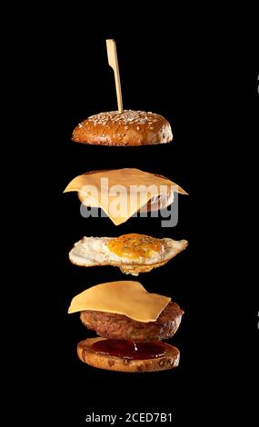 ingrédients de cheeseburger volant : côtelette de viande, pain de graines de sésame, fromage, œuf frit et ketchup de tomate, fond noir Banque D'Images