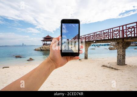 Rognez la main à l'aide d'un smartphone et prenez des photos de la passerelle sur la côte tropicale de l'île de Wuzhizhou, Hainan, Chine Banque D'Images