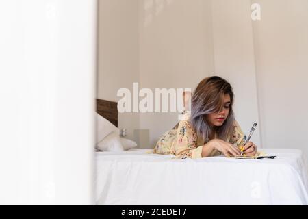 Belle jeune femme allongé sur un lit confortable et écrivant dans un petit carnet avec main gauche tout en se reposant dans une pièce élégante Banque D'Images