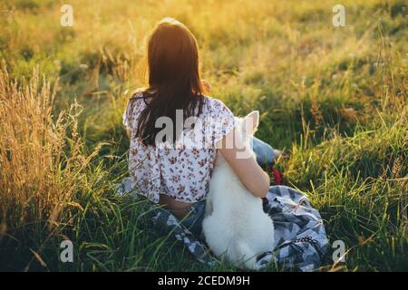 Femme assise avec un chiot blanc mignon et regardant le coucher de soleil dans la prairie d'été. Une jeune fille élégante se détendant avec son adorable chiot moelleux dans une lumière chaude au coucher du soleil. Banque D'Images