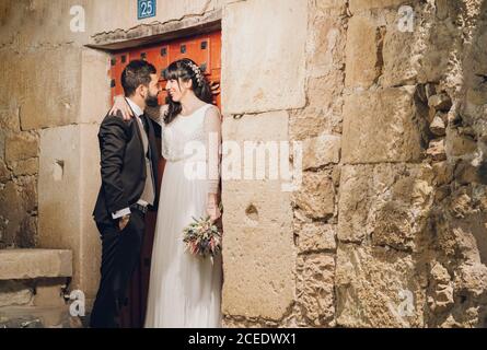 Bonne belle mariée dans la robe de mariage tenant le bouquet et le marié en costume noir souriant et s'embrassant les uns les autres près de la pierre mur Banque D'Images