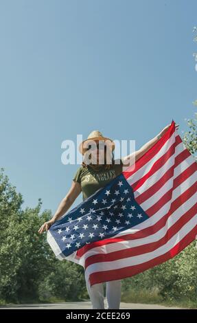 Bonne femme marchant avec un drapeau américain et célébrant sur une route solitaire. Journée spéciale pour célébrer le 4th juillet Banque D'Images