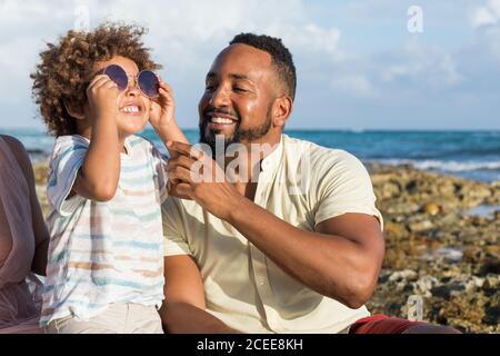 Joyeux homme ethnique adulte donnant des lunettes de soleil pour essayer de peu fils à l'océan Banque D'Images