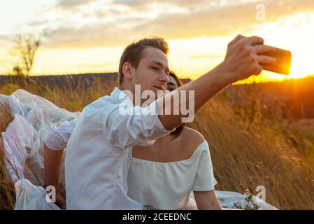 jeune couple un gars et une fille de la génération z, faire une photo de selfie, assis dans l'herbe dans le champ, sur la toile de fond du soleil couchant. été Banque D'Images