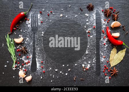 Diverses herbes et épices sur table en pierre sombre. La silhouette des couverts sur une table noire est accompagnée de sel, d'épices, de piment, de romarin et d'ail. Banque D'Images