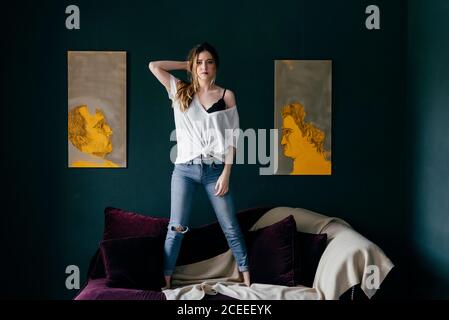 Belle jeune femme dans un ensemble élégant regardant l'appareil photo tout en se tenant sur un canapé doux dans une pièce confortable Banque D'Images