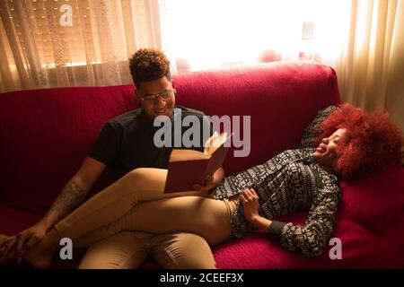 Jeune couple afro-américain élégant qui se câlin sur un canapé rouge et lit réservez à haute voix pour passer du temps ensemble Banque D'Images