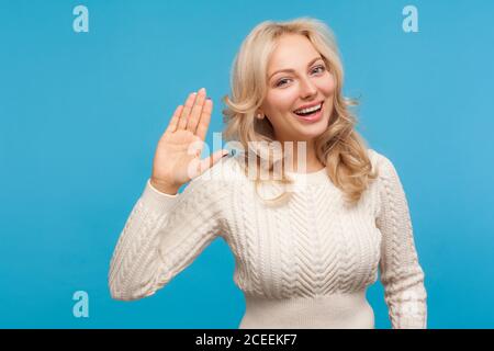 Sympathique heureuse femme blonde montrant la main levée en disant salut avec le sourire crasseux sur le visage, salutation optimiste. Prise de vue en studio isolée sur fond bleu Banque D'Images
