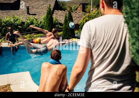 les hommes méconnaissables s'amusent et sautent à la piscine tout en jouant au ballon. Banque D'Images