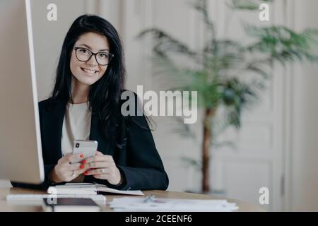 Une employée de bureau heureuse dans une élégante veste noire parcourt son smartphone avec une expression gaie, envoie des messages texte, travaille sur un projet important, pose Banque D'Images