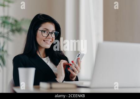 Prise de vue horizontale des messages européens de la femme brunette heureuse sur smartphone moderne, donne une consultation en ligne, sourit positivement, porte des lunettes et pour Banque D'Images