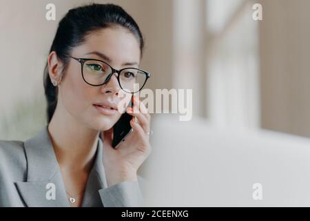 Gros plan d'une agréable brunette européenne femme porte des lunettes transparentes, focalisées sur l'écran d'ordinateur portable, a la conversation téléphonique pendant la remot Banque D'Images