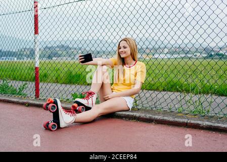 Jeune fille portant des patins à roulettes et montrant deux doigts à l'appareil photo du smartphone prenant selfie dans la rue. Banque D'Images
