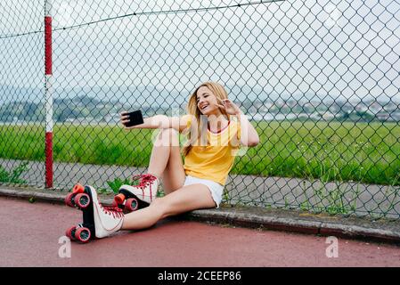 Jeune fille portant des patins à roulettes et montrant deux doigts à l'appareil photo du smartphone prenant selfie dans la rue. Banque D'Images